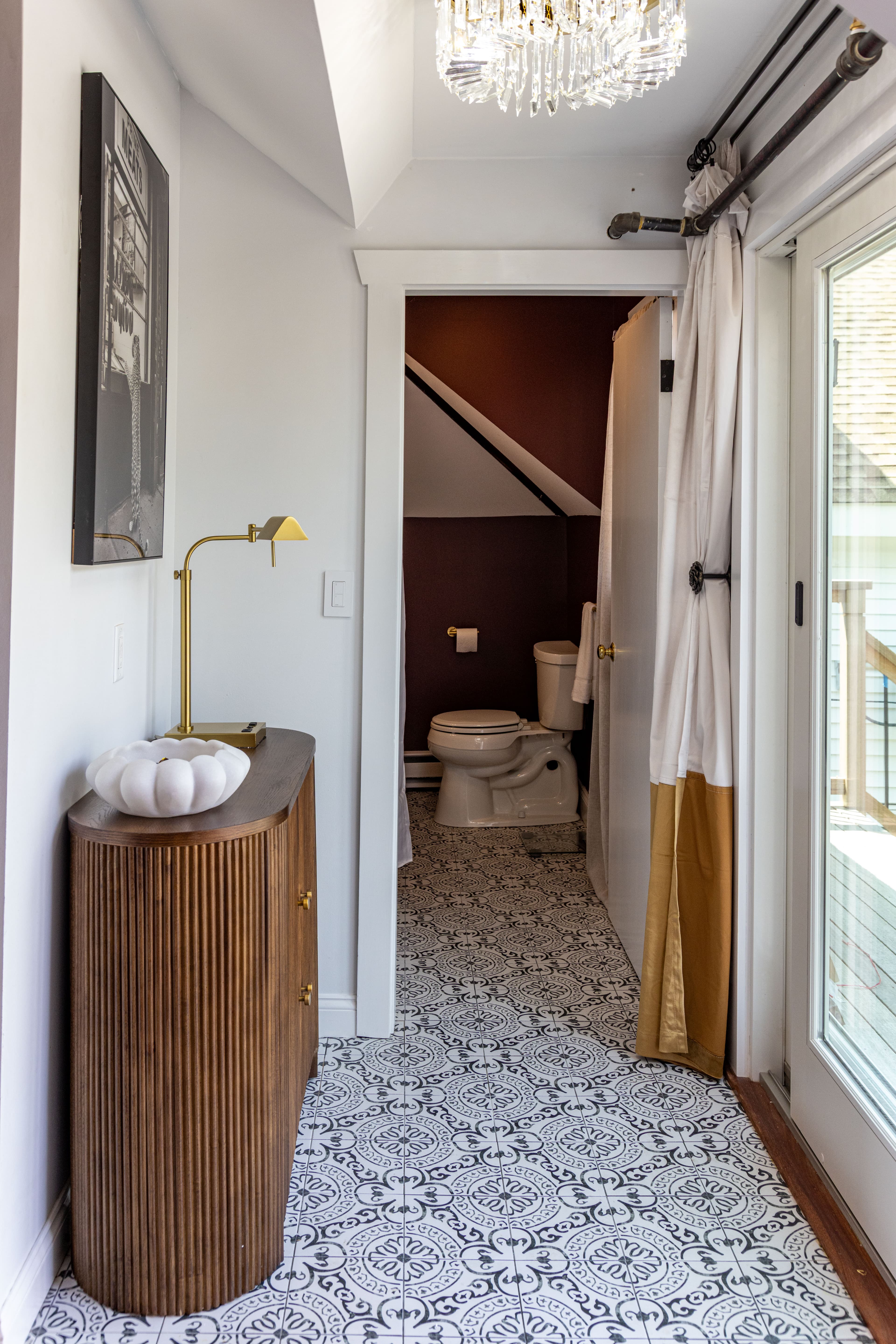 Interior view of a hallway leading to a bathroom, featuring a wooden console, patterned tile floor, and a chandelier.