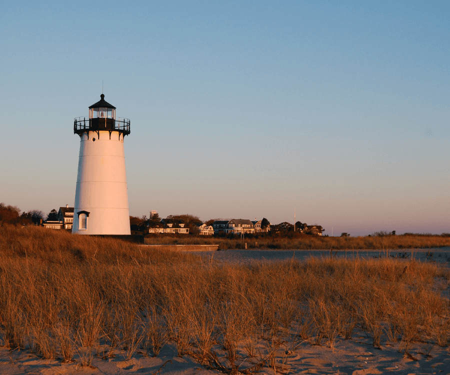 A white lighthouse stands beside sandy grass dunes at sunset, with buildings in the background.