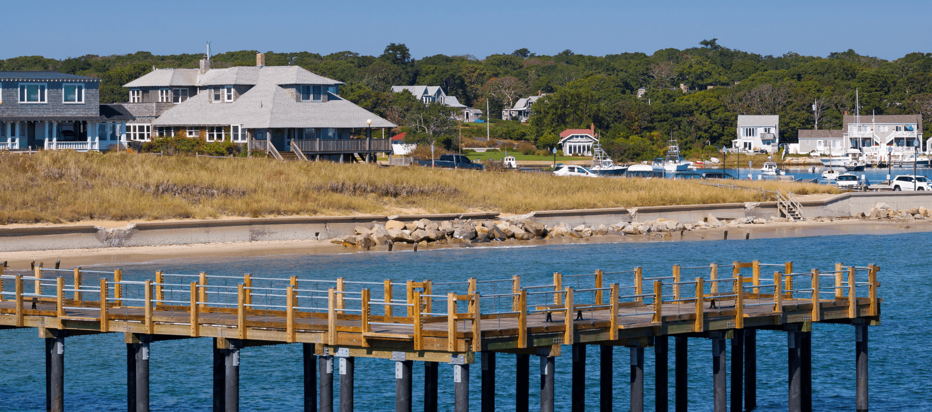 A wooden pier extends into calm water with grassy shores and houses in the background.