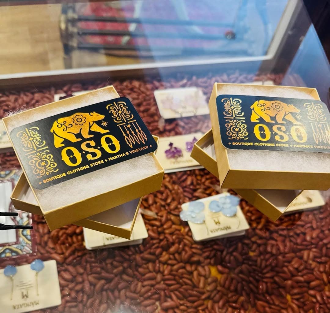 Two open boxes displaying the "OSO" logo, surrounded by colorful earrings and beans in a display case.