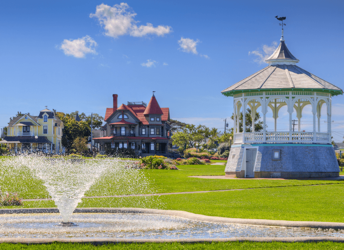 A gazebo stands beside a fountain and historic homes in a sunny park.