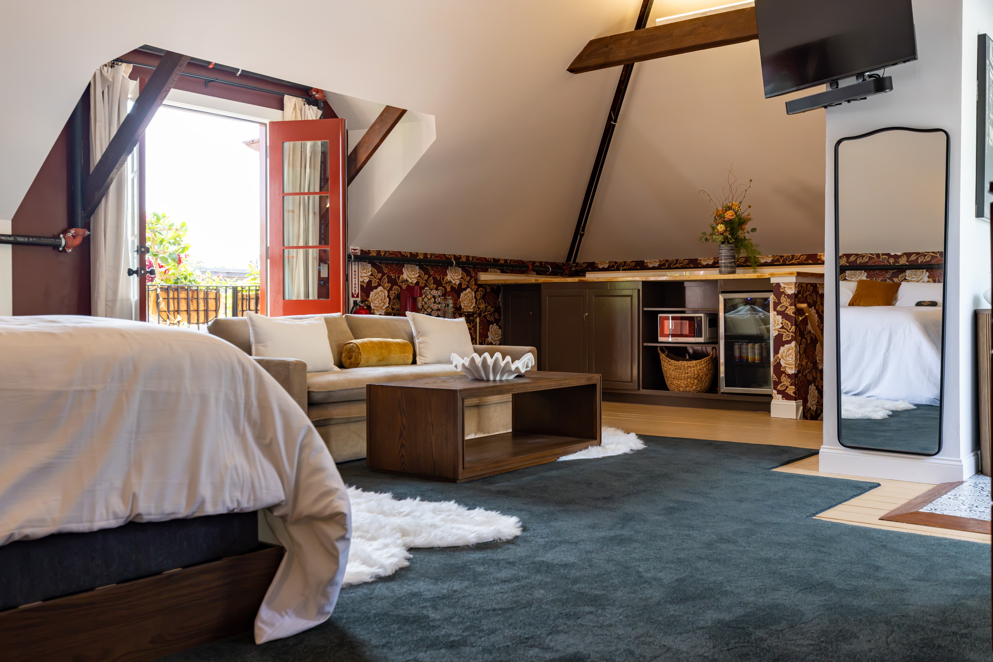 Cozy attic bedroom with a bed, seating area, kitchenette, and open balcony doors.