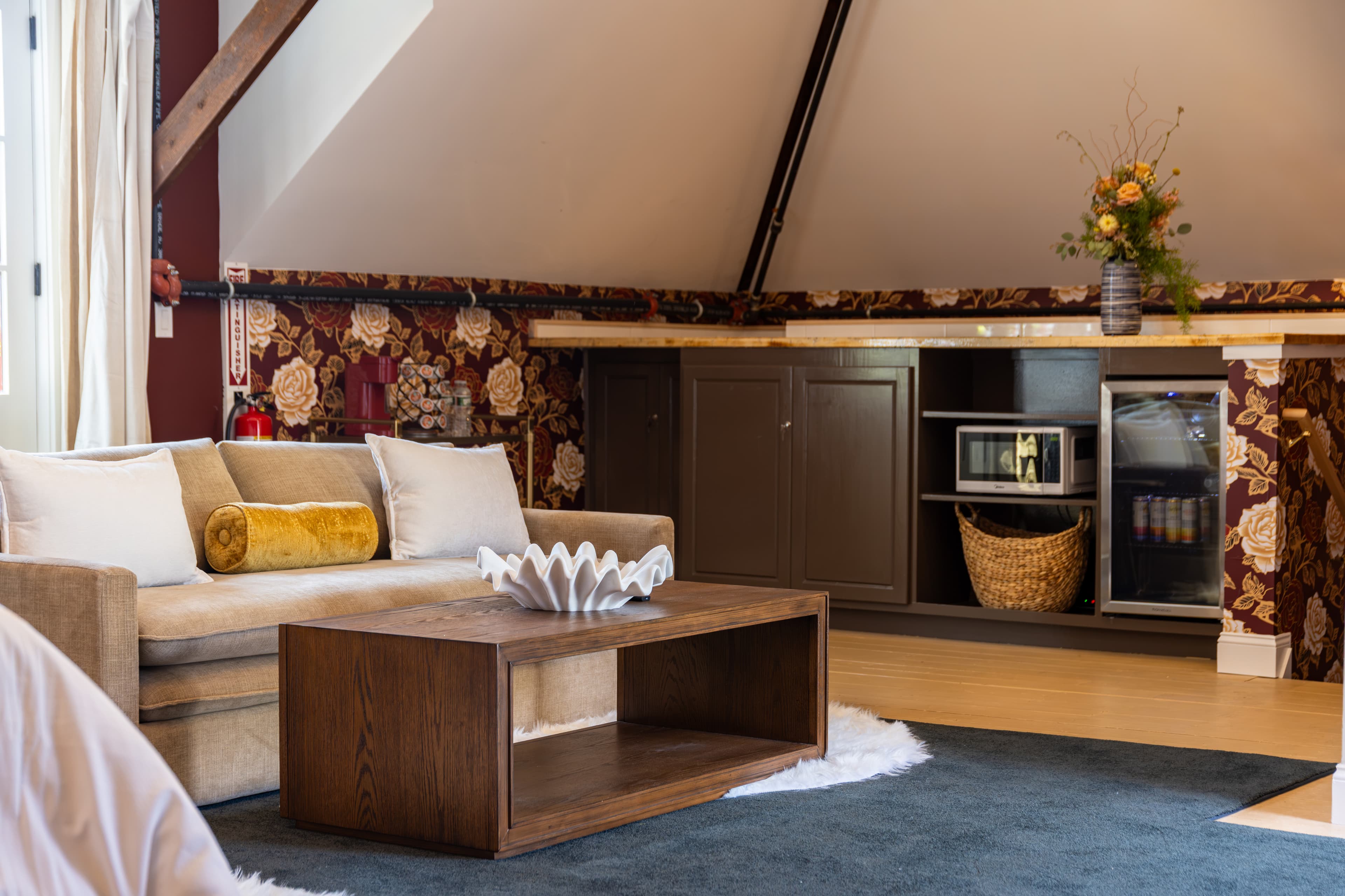 A cozy living space featuring a beige couch, wooden coffee table, and floral wallpaper with a kitchenette in the background.