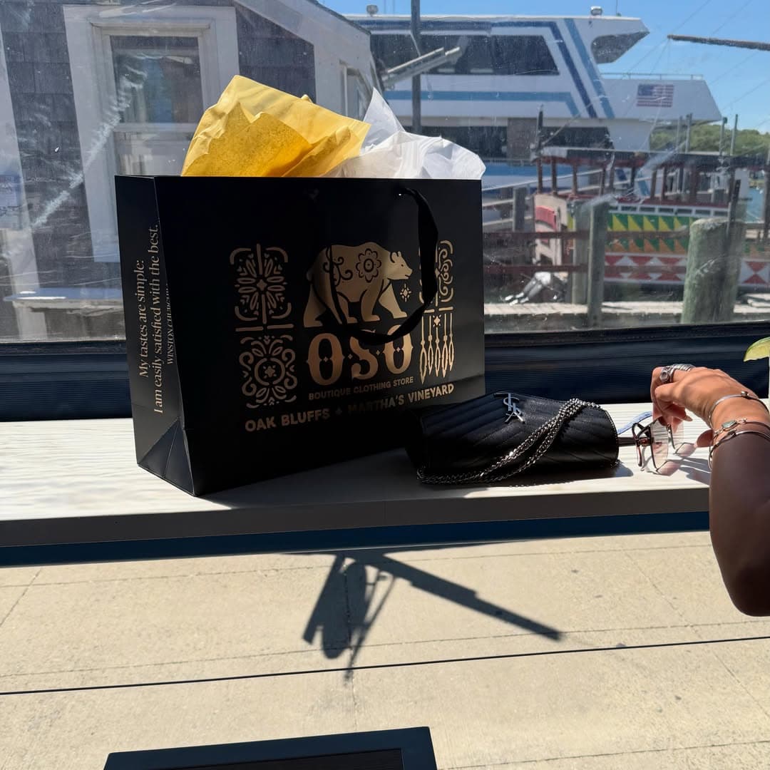 A black shopping bag with the logo "OSU" and tissue paper sits on a table beside a hand holding sunglasses.